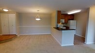 This single-floor space presents a clean and functional open-concept layout that seamlessly connects the living, dining, and kitchen areas. The living and dining area is generously proportioned, featuring light neutral-toned carpet flooring and warm beige walls accented by white baseboards and a classic chair rail molding that runs along the perimeter, adding a touch of traditional detail to the interior. A pendant light fixture hangs centrally over the dining area, providing focused overhead illumination and serving as a subtle decorative focal point.

The kitchen is positioned at the far end of the open space and is defined by a raised breakfast bar with a dark countertop, offering a natural visual and functional divider between the cooking and living areas. The kitchen features warm-toned wood cabinetry, a white refrigerator, a range with an overhead microwave, and under-cabinet lighting that brightens the workspace. The layout is efficient and well-organized, making the most of the available space.

Overall, the space conveys a comfortable, well-maintained ambiance with a straightforward design that emphasizes practicality and ease of living. The consistent use of neutral tones throughout creates a cohesive and inviting atmosphere.
