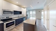 Located at 827 Webster St in Fort Wayne, Indiana, this thoughtfully designed unit offers a contemporary open-concept layout with efficient use of space across a single floor. The kitchen is a standout feature, showcasing a two-tone design with sleek white upper cabinetry and dark lower cabinets, granite countertops, and a full suite of stainless steel appliances including a French door refrigerator, electric range, built-in microwave, and dishwasher. A central island with a matching granite surface provides additional prep space and casual seating. Pendant lighting and recessed fixtures illuminate the kitchen, which flows seamlessly into the living area. Wide-plank hardwood-style flooring runs throughout the main living spaces, adding warmth and continuity. A large sliding glass door leads to a private balcony, flooding the interior with natural light and offering open views of the surrounding Fort Wayne cityscape. The bathroom features a full tub-shower combination with clean tile flooring and granite-topped vanity. A dedicated in-unit laundry area accommodates a full-size washer and dryer with overhead wire shelving for added storage. The overall aesthetic is clean, modern, and move-in ready.