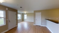 This single-floor space presents a clean, functional design highlighted by light hardwood flooring that runs throughout, lending warmth and continuity to the interior. The open main area is finished with neutral, earthy-toned walls that create a calm and versatile backdrop suitable for a variety of uses. White panel doors provide access to adjoining rooms or storage spaces, contributing to the layout's flexibility. A centrally positioned flush-mount light fixture illuminates the space, while a double window along one wall allows natural light to filter in, brightening the interior and offering a view to the outdoors. White baseboards frame the perimeter of the room, adding a crisp contrast against the warm-toned walls and flooring. A staircase railing is partially visible near one edge of the space, suggesting additional vertical access nearby. Ceiling-mounted ventilation ensures practical comfort throughout. The overall ambiance is understated and move-in ready, with a straightforward layout that provides ample room to personalize and arrange as needed.