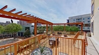 This rooftop terrace is a generously sized outdoor space designed for both social gatherings and quiet relaxation, elevated above a vibrant urban streetscape. The terrace's most prominent architectural feature is its series of bold, richly stained wood pergolas, which provide partial shade while framing open views of the surrounding cityscape, including nearby buildings, mature trees, and a lively street-level environment below.

The terrace is finished with warm-toned tile and flagstone flooring throughout, adding a refined, cohesive aesthetic to the space. Multiple distinct seating zones are thoughtfully arranged across the terrace. A large rectangular dining table with chairs accommodates group meals or meetings, while a separate lounge area features slatted wood furniture arranged around a fire pit, offering a more relaxed setting for conversation. Additional benches and accent planters with ornamental grasses contribute to the inviting atmosphere.

Perimeter railing in a matching warm wood tone defines the edges of the terrace, ensuring both safety and unobstructed views. The open-sky orientation allows for abundant natural light throughout the day, and the urban backdrop provides dynamic visual interest from every vantage point, making this terrace a standout feature of the overall space.