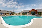 Located at 200 Travis Dr in McDonough, GA, this generously sized outdoor amenity space serves as the centerpiece of a modern residential community. Spanning a substantial single-level footprint, the area is anchored by a large, freeform resort-style swimming pool with crystal-clear turquoise water and a smooth curved perimeter. The expansive pool deck is finished in textured aggregate concrete, providing both aesthetic appeal and slip resistance, and is furnished with a generous array of chaise lounges, chairs, and shaded umbrella seating for relaxation.

The adjacent clubhouse is an architectural highlight, featuring a striking gabled roofline with exposed timber framing, stone and fiber cement exterior cladding, and floor-to-ceiling windows that blur the line between indoor and outdoor spaces. A covered patio extends from the clubhouse directly toward the pool, outfitted with comfortable lounge furniture for year-round use.

Surrounding the pool deck, additional amenities include hammock stations, a pergola-covered cabana area, and landscaped garden beds with decorative gravel accents. A wrought iron perimeter fence defines the space while maintaining open sightlines to the multi-story residential buildings beyond. The overall ambiance is that of a well-designed, resort-caliber outdoor retreat suited for both leisurely relaxation and social gatherings.