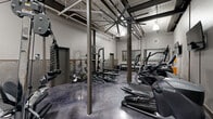 Located at 710 Greenville St in LaGrange, Georgia, this fitness facility offers a thoughtfully designed exercise space with an industrial aesthetic. The room features soaring ceilings with exposed ductwork, structural steel columns, and pendant lighting that create an open, warehouse-inspired atmosphere. The polished concrete floors in a deep charcoal finish add durability and a modern edge to the space.

The facility is comprehensively equipped for a full-body workout experience. The cardio zone features multiple TRUE-brand treadmills, ellipticals, stair climbers, and stationary bikes, each outfitted with individual display screens. The strength training area includes a robust power rack loaded with weight plates, a multi-station cable machine, adjustable benches, and a full dumbbell rack ranging across multiple weight increments. Kettlebells and medicine balls round out the free weights section.

Exposed brick walls painted in light gray lend a classic industrial character, while large ceiling fans provide ventilation throughout. A pass-through window connects to an adjacent area, and a dedicated restroom is accessible directly from the gym floor. Mounted flat-screen televisions enhance the workout environment. The overall ambiance is functional, clean, and purposefully designed for serious fitness use.