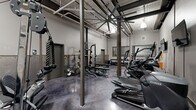 Located at 710 Greenville St in LaGrange, Georgia, this fitness facility offers a thoughtfully designed exercise space with an industrial aesthetic. The room features soaring ceilings with exposed ductwork, structural steel columns, and pendant lighting that create an open, warehouse-inspired atmosphere. The polished concrete floors in a deep charcoal finish add durability and a modern edge to the space.

The facility is comprehensively equipped for a full-body workout experience. The cardio zone features multiple TRUE-brand treadmills, ellipticals, stair climbers, and stationary bikes, each outfitted with individual display screens. The strength training area includes a robust power rack loaded with weight plates, a multi-station cable machine, adjustable benches, and a full dumbbell rack ranging across multiple weight increments. Kettlebells and medicine balls round out the free weights section.

Exposed brick walls painted in light gray lend a classic industrial character, while large ceiling fans provide ventilation throughout. A pass-through window connects to an adjacent area, and a dedicated restroom is accessible directly from the gym floor. Mounted flat-screen televisions enhance the workout environment. The overall ambiance is functional, clean, and purposefully designed for serious fitness use.