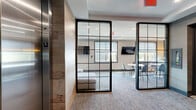 Located on the 5th floor at 975 Bonnel Ct in Union, NJ, this thoughtfully designed lounge space offers a refined and contemporary communal environment. The entry foyer sets an elegant tone with textured gray wall panels, plush carpet flooring, and a statement console table adorned with sculptural décor beneath a large round mirror — all positioned adjacent to elevator access.

Floor-to-ceiling steel-framed glass partitions divide the space into distinct functional zones while maintaining an open, transparent feel throughout. The living area features a generously sized sectional sofa with vibrant accent pillows, round upholstered ottomans, and decorative gold wall art set against soft gray walls with a tray ceiling detail. Natural light pours in through large windows, enhancing the airy ambiance.

A separate seating zone accommodates a marble-top round table with modern chairs and multiple wall-mounted flat-screen televisions, making it well-suited for both casual gatherings and media viewing. Colorful framed artwork adds personality throughout the space. Recessed lighting complements the natural light, ensuring a well-lit environment at all times. The overall design balances modern sophistication with a welcoming, comfortable atmosphere.
