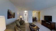 This single-floor residence offers a practical and comfortable layout designed for everyday living. The living area is carpeted and features neutral-toned walls that create a calm, understated ambiance. A cushioned sofa and coffee table anchor the space, while wall art adds a touch of personality. The room flows seamlessly into the dining area, which is defined by a ceiling fan with a light fixture overhead and accommodates a dining table and chairs, making it well-suited for both casual meals and gatherings. Adjacent to the living and dining spaces, the kitchen is accessible via an open passageway and features a stainless steel refrigerator, light-toned flooring, and functional cabinetry. An intercom panel is mounted on the wall near the transition between rooms, adding a practical touch. The overall layout promotes an easy flow between spaces, and the consistent use of neutral colors and carpeted flooring throughout gives the unit a cohesive, unified feel. Natural and artificial lighting work together to keep the interiors feeling bright and welcoming across all areas of the home.