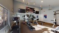 Located at 1300 E Fort Lowell Rd in Tucson, Arizona, this fitness center at College Town Tucson is a purpose-built, single-floor workout facility designed to meet the exercise needs of its residents. The space features soaring vaulted ceilings that create an open, airy environment, complemented by recessed lighting and ceiling fans that maintain comfort during workouts. The flooring consists of durable, patterned carpet tiles throughout, providing a functional surface suited for a variety of exercises. Natural light streams in through multiple windows, some featuring decorative etched glass with a wildcat motif, offering views of the surrounding outdoor landscape including desert vegetation. The facility is well-stocked with a comprehensive range of equipment, including treadmills, an elliptical trainer, a stationary bike, a multi-station cable weight machine, adjustable benches, free weights, and a medicine ball. Large wall-mounted flat-screen televisions are positioned throughout the space for entertainment during workouts. A full-length mirror along one wall enhances the sense of space and allows users to monitor their form. The walls are adorned with University of Arizona Wildcats-themed signage and motivational sports imagery, reinforcing the college community atmosphere. Posted fitness center rules ensure a safe and organized environment for all users.
