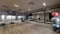 This contemporary fitness facility is thoughtfully designed to support a wide range of workout activities within a single, open-plan floor. The studio's most striking feature is the expansive wall of floor-to-ceiling mirrors framed in dark trim, which line the main workout area and create a sense of depth and openness throughout the space. The light-toned, hardwood-style flooring provides a clean, durable surface ideal for both cardio and strength training activities. Recessed and flush-mounted ceiling lights ensure the entire area is evenly and brightly illuminated, contributing to an energetic yet focused atmosphere. Glass-paneled partitions divide workout zones while maintaining visual connectivity across the studio, allowing natural light to filter through the space. Various fitness equipment is visible throughout, including medicine balls, step platforms, resistance training tools, and interactive fitness screens, catering to diverse training needs. A mounted flat-screen television adds a multimedia element to the environment. The overall design balances functionality with a sleek, modern aesthetic, making this fitness studio a well-equipped and visually inviting space for exercise and wellness activities.