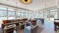This stylish single-floor amenity space is designed for both relaxation and recreation, offering a thoughtfully curated layout that blends comfort with entertainment. The interior is anchored by expansive floor-to-ceiling windows that wrap around the room, flooding the space with natural light and providing panoramic views of the surrounding wooded landscape and outdoor pool area beyond.

The main lounge area features generous sectional seating in neutral tones, accented with bold orange throw pillows, arranged around a pair of wooden coffee tables atop a textured area rug. A striking stacked-stone fireplace with a modern linear firebox serves as a focal point on one wall, topped by a wall-mounted flat-screen television. An eye-catching orange upholstered bench with bold artwork adds a vibrant pop of color near the entryway.

The game room section is equipped with a rustic wood-toned billiards table and a full shuffleboard table, catering to a range of recreational interests. An additional wall-mounted TV overlooks the game area, making it ideal for social gatherings.

Underfoot, a distinctive herringbone-patterned hardwood floor runs throughout, adding warmth and visual sophistication. Overhead, a combination of drum pendant lights and a decorative ceiling fixture with a gold interior provides layered ambient lighting, completing the polished yet inviting atmosphere of this community gathering space.