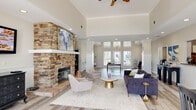 Located at 1530 Wimbledon Dr in Greenville, NC, this single-floor community amenity space combines comfort and functionality in a thoughtfully designed layout. The lounge area immediately impresses with soaring vaulted ceilings, crown molding, and light hardwood-style flooring that runs throughout. A dramatic floor-to-ceiling stacked stone fireplace serves as the centerpiece, topped with a white mantel and framed artwork that adds a refined touch. The space is furnished with plush seating, a velvet sofa, and a gold accent coffee table, all anchored by a neutral area rug. Recessed lighting and a ceiling fan complement the abundant natural light streaming through large windows and glass doors. An adjacent dining nook features wainscoting, multiple windows with outdoor views, and a raised dining table — ideal for casual gatherings. The fitness center is generously appointed with cardio machines, a multi-station weight machine, a full dumbbell rack, medicine balls, and wall-mounted mirrors. Its bold lime green accent stripe and checkered flooring in the cardio area give the gym an energetic atmosphere, while natural light from multiple windows keeps the environment bright and motivating.