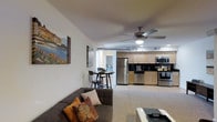 This single-floor residence offers a thoughtfully designed open-concept layout that combines the living area, dining nook, and kitchen into one cohesive, functional space. The living area features plush carpeted flooring and is anchored by a comfortable seating arrangement, with a ceiling fan overhead providing both light and airflow. Recessed lighting complements the clean white walls, which are accented by colorful wall art, lending warmth and personality to the space.

The kitchen is positioned at the rear of the open floor plan and is well-appointed with a full suite of stainless steel appliances, including a refrigerator, microwave, and range. Light wood cabinetry extends to the upper and lower sections, paired with dark countertops for a modern contrast. Exposed ductwork along the ceiling adds an industrial touch while maintaining the open, airy feel of the space.

Adjacent to the kitchen, a compact bistro-style dining nook with bar-height seating provides an intimate spot for casual meals. A hallway visible beyond the dining area leads to additional rooms, suggesting a well-organized floor plan. Overall, the space presents a clean, modern aesthetic with practical design choices that maximize both comfort and functionality throughout the single level.