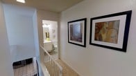 This single-level space is thoughtfully laid out with a welcoming interior design defined by neutral wall tones, textured ceilings, and warm, understated finishes. The upper hallway serves as a central connector, featuring plush carpeting underfoot and a classic white-railed staircase that leads down to a tiled lower landing. The hallway walls are adorned with a pair of bold, framed abstract artworks, adding a decorative focal point to an otherwise clean and minimal corridor. Recessed ceiling lighting provides consistent illumination throughout the passage. Just off the hallway, a full bathroom is accessible and well-appointed, showcasing tile flooring, a white vanity with mirror and overhead lighting, and a standard toilet — all maintained in a bright, clean aesthetic. The lower landing, visible from the hallway above, features tile flooring and hints at additional living areas beyond. Throughout the space, the combination of carpet, tile, and painted surfaces creates a cohesive and functional environment. The layout emphasizes efficient use of space while maintaining a tidy, livable atmosphere suitable for everyday comfort.