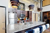 Located at 1101 Hawthorne Lane in Charlotte's vibrant Plaza Midwood neighborhood, this one-bedroom, one-bathroom loft showcases an impressive industrial-chic aesthetic rooted in its converted warehouse origins. The open-plan living area commands attention with soaring ceilings, exposed brick walls, and a dramatic arched window that floods the space with abundant natural light. Polished concrete floors run throughout, complementing the raw architectural character. The kitchen features granite countertops and a breakfast bar with seating, seamlessly connecting to the living area. The spacious bedroom continues the loft's bold design language, with exposed brick, floor-to-ceiling steel-framed windows, and direct access to a private outdoor patio — an uncommon amenity that provides a serene retreat within the urban setting. The patio itself is framed by the original brick walls of the surrounding building, offering a sheltered and intimate outdoor space. The bathroom is richly appointed with a granite vanity, dark cabinetry, decorative wall-to-ceiling gallery arrangements, and warm sconce lighting that creates an atmospheric, boutique-hotel feel. Throughout the space, high ceilings and large industrial-style windows reinforce a sense of openness and volume that far exceeds the physical footprint of this thoughtfully designed urban loft.
