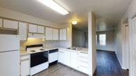 This single-story space features a practical, well-organized kitchen as its central highlight. The kitchen is outfitted with white cabinetry that extends to the upper walls, providing ample storage, along with a built-in electric range with cooktop and a dishwasher. A double-basin stainless steel sink is situated along the countertop, which wraps in an L-shaped configuration to maximize workspace. Under-cabinet lighting above the range adds functional illumination, complemented by a flush-mount ceiling fixture. The textured ceiling and neutral-toned walls create a clean, understated backdrop throughout.

Wood-style flooring runs consistently through the kitchen and into the adjoining areas, lending a cohesive feel across the layout. A half-wall partition separates the kitchen from a neighboring room, maintaining an open sightline while defining each space. Through the opening, an additional room is visible with a window that allows natural light to filter in, brightening the overall interior. The single-floor layout ensures easy navigation between spaces, making the unit feel connected and accessible throughout.