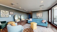 Baker Social is a thoughtfully designed social lounge located in the Baker neighborhood of Denver, Colorado. The interior is defined by a sophisticated yet eclectic aesthetic, featuring slate-blue walls accented by crisp crown molding and warm brick-patterned herringbone flooring that anchors the entire space. Seating is an artful mix of styles, including a tufted blue chesterfield sofa, ornate high-backed dome chairs upholstered in turquoise floral fabric, a rich emerald green armchair, and swivel lounge chairs in muted tones — all complemented by yellow leather ottomans that add a vibrant pop of color. Recessed lighting and floor lamps provide a warm, ambient glow throughout. One wall features rich wood paneling surrounding a mounted flat-screen television, creating a dedicated media nook with built-in cabinetry. Curated botanical and wildlife artwork decorates the walls, reinforcing the space's distinctive character. Large floor-to-ceiling glass doors and windows lead directly to a covered outdoor terrace, which offers a generous bar-height counter with blue metal stools, Adirondack seating, and sweeping panoramic views of the Denver cityscape and surrounding treetops — making it an ideal setting for gatherings and social events.