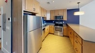 This single-floor unit blends industrial character with modern finishes throughout its well-organized layout. The kitchen serves as a central highlight, featuring light maple cabinetry, stainless steel appliances including a full-size refrigerator, microwave, and electric range, complemented by gray laminate countertops and a bold navy blue accent wall. Tile flooring in a warm yellow-beige tone adds a clean, practical surface underfoot. A peninsula counter with a pendant light overhead connects the kitchen to the adjacent living area, creating an open, functional flow between spaces.

The living area transitions seamlessly from the kitchen and is carpeted in a neutral gray tone, providing a comfortable, understated backdrop. Exposed original brick walls and tall, black-framed industrial windows are defining architectural features, allowing natural light to pour in while offering views of the surrounding urban streetscape. The soaring ceiling height in this area reinforces the building's historic character.

An additional carpeted room with smooth gray walls and recessed lighting provides a versatile, quiet space suitable for a bedroom or office. The bathroom is finished with large-format tile flooring, a wood-toned vanity with a white sink, a wall-mounted mirror, and a glass-enclosed shower, combining practical function with a clean, contemporary aesthetic.