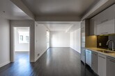 This contemporary single-floor residence showcases a thoughtfully designed open-concept layout that seamlessly connects the kitchen and living areas. Rich, dark hardwood flooring extends throughout the main living spaces, providing a striking contrast against the clean white walls and ceilings with architectural beam details.

The kitchen is a standout feature, outfitted with flat-front grey cabinetry that reaches toward the ceiling for ample storage, a vibrant yellow countertop and backsplash with reflective dark paneling, a gas range with oven, an under-cabinet range hood, and a stainless steel sink. The bold color palette gives the kitchen a modern, energetic character.

The expansive living area is bathed in natural light, offering a generous, versatile space with a clean, minimalist aesthetic. A bedroom features a large window framing views of the surrounding urban streetscape, allowing abundant daylight to fill the room.

The bathroom is finished with a white floating vanity topped with a large mirror, dark mosaic tile flooring, and a tiled shower-tub combination, creating a clean and functional space. Throughout the unit, the consistent use of white walls, dark flooring, and modern fixtures contributes to a cohesive, contemporary aesthetic well-suited for modern living.