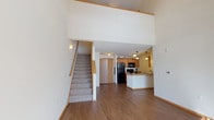 This two-story residence is defined by its dramatic vaulted ceilings on the main level, creating an expansive, airy atmosphere from the moment you enter. The open main floor showcases rich hardwood-style flooring that flows throughout the living area, which benefits from a sliding glass door that draws in abundant natural light and provides outdoor views.

The kitchen is thoughtfully designed with warm honey-toned cabinetry extending to the ceiling, a raised breakfast bar peninsula, pendant lighting, and black appliances including a refrigerator, range, and built-in microwave. A double sink with additional counter space adds functional workspace to the layout.

Carpeted stairs with a wood handrail lead to the upper level, where an open loft overlooks the main floor below. This flexible upper space features plush carpeting and a ceiling fan, making it suitable for a variety of uses. Additional bedrooms on this level are carpeted and bright, each equipped with ceiling fans and windows that allow natural light in.

The bathrooms include a full bath with a double-sink vanity, warm wood cabinetry, and a glass-enclosed shower, as well as a secondary bath with tile flooring and a shower-tub combination. A dedicated laundry area with a full-size washer and dryer and overhead shelving adds everyday convenience.