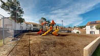 Located at 36523 25th St E in Palmdale, California, this outdoor community amenity space serves as a central recreational hub for the surrounding residential complex. The area is thoughtfully divided into distinct zones, each designed to accommodate different leisure activities.

The playground area is a standout feature, showcasing a large, colorful multi-structure play system with multiple slides — including both straight and spiral designs — climbing elements, and tower structures topped with vibrant orange and red canopy roofs. The play surface is covered with wood chip mulch, providing a safe and cushioned ground cover. Seating benches are positioned nearby for supervision and rest.

Adjacent to the playground, a generous open lawn area features well-maintained artificial turf, offering a versatile space for outdoor activities. A wide concrete walkway borders the lawn, providing clean pedestrian access throughout the space.

A fenced swimming pool is situated at one end of the complex, accompanied by what appears to be a pool house or clubhouse structure with arched architectural detailing and a tile roof, consistent with the Spanish-influenced design of the surrounding residential buildings. The complex buildings themselves feature white stucco exteriors, red tile roofs, and solar panels on select units. The entire amenity area is enclosed by chain-link fencing, with mature trees and open sky contributing to a bright, open atmosphere.