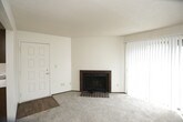This single-floor unit offers a functional and straightforward layout with several notable features throughout. The living area is carpeted and anchored by a brick fireplace with a metal surround, adding a classic focal point to the space. A ceiling fan overhead provides practical comfort, while an open passageway connects the living room directly to the kitchen. A sliding glass door in an adjacent room floods the space with natural light through vertical blinds, creating a bright and open atmosphere.

The galley-style kitchen is equipped with dark wood cabinetry above and below white laminate countertops, offering ample storage. White appliances, including a refrigerator, electric range, and dishwasher, line one wall, while a stainless steel sink is positioned along the opposite counter. Wood-look flooring runs through the kitchen, adding warmth to the space.

The bedroom is generously sized with plush carpeting, a textured ceiling, a window with a view to the outside, and access to a walk-in closet and a private bathroom. The bathroom features wood-look flooring, a full tub-shower combination, a toilet, and a vanity with a wood-framed mirror and integrated sink.