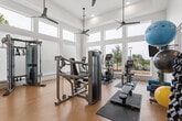 Located at 4770 Military Drive West in San Antonio, Texas, this fitness center is a thoughtfully designed exercise facility that offers a comprehensive workout experience within a bright and welcoming space. The room features soaring ceilings with large, multi-pane windows that span the walls, flooding the interior with abundant natural light and offering views of the surrounding outdoor environment. Ceiling fans are strategically placed overhead to ensure proper air circulation throughout workouts.

The warm-toned hardwood flooring provides a clean and durable surface underfoot, complementing the crisp white walls and modern aesthetic. The facility is outfitted with a robust selection of Matrix-brand equipment, including treadmills, an elliptical trainer, a cable crossover machine, and a seated press station, catering to both cardiovascular and strength training needs. A dedicated functional training area features a well-organized rack stocked with medicine balls, resistance bands, stability balls, and foam rollers, alongside a flat bench for versatile exercises.

A standout design feature is an eye-catching accent wall clad in multi-toned blue and grey stacked tile, set above a built-in storage unit that keeps towels and fitness accessories neatly organized. A compact refrigerator is also available for added convenience, rounding out this fully appointed and visually striking fitness space.