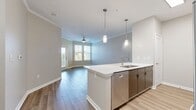 This contemporary 750 square foot apartment offers a thoughtfully designed living space with a blend of style and functionality. The open-concept layout seamlessly integrates the kitchen, living, and dining areas, creating an expansive feel. The kitchen boasts sleek white cabinetry, contrasting with warm wood-tone lower cabinets, and is equipped with stainless steel appliances including a refrigerator, stove, and built-in microwave. A large kitchen island with a white countertop provides additional workspace and casual dining options.

The spacious primary bedroom, measuring 210 square feet, features plush carpeting, a ceiling fan, and large windows that flood the room with natural light. The bathroom is modern and well-appointed with a single vanity featuring ample storage, a large mirror, and a toilet. Light-colored tile flooring complements the overall neutral palette.

Throughout the apartment, wood-look flooring adds warmth and durability to the main living areas. Crown molding and baseboards provide elegant finishing touches. The unit also includes a walk-in closet with wire shelving for organization. Strategically placed windows with white blinds allow for privacy while maximizing natural light, creating a bright and airy atmosphere throughout the space.