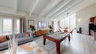 Located at 1313 Stone Mill Way in Stone Mountain, Georgia, this thoughtfully designed single-floor space blends modern style with functional versatility. The main living and entertainment area is the heart of the space, boasting striking white-painted exposed wood beam ceilings with shiplap detailing that lend a warm, architectural character. Light grey hardwood-style flooring runs throughout, complementing the neutral tones of the interior. Large floor-to-ceiling windows and glass doors flood the room with natural light while offering serene views of the surrounding wooded landscape, creating an open, airy ambiance. The entertainment area features a full-size pool table as a centerpiece, alongside comfortable tufted grey sofas accented with warm terracotta pillows, a wall-mounted flat-screen TV, and a ceiling fan for year-round comfort. High walls with transom-style windows add verticality and architectural interest to the space. A well-appointed kitchenette is also present, featuring rich cherry wood cabinetry, dark granite countertops, a tile backsplash, stainless steel appliances including a refrigerator and dishwasher, and ample storage. The overall atmosphere is sophisticated yet inviting, making this space well-suited for gatherings, entertainment, or relaxed everyday use.