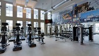 Located at 1 Kennedy Ave in Danbury, Connecticut, this single-floor fitness center is a well-equipped and visually striking space designed to support a full range of workout routines. The facility boasts impressively high ceilings with exposed ductwork and industrial-style ceiling fans, lending the space an open, airy, and modern aesthetic. Rows of large windows line the walls, flooding the interior with abundant natural light and offering views of the surrounding neighborhood.

The cardio area is outfitted with a comprehensive selection of machines, including treadmills, ellipticals, stationary bikes, and rowing machines, all positioned to take advantage of the natural light and views. The strength training section features a cable/pulley machine, a full rack of dumbbells, weight benches, and additional resistance equipment, providing everything needed for a complete strength workout. Mounted televisions are conveniently placed throughout the space for entertainment during workouts.

A standout design element is the large, vibrant mural celebrating the historic Great Danbury State Fair, which adds a strong sense of local character and community identity to the space. The clean, light-colored flooring and well-organized layout contribute to a welcoming and functional environment suitable for all fitness levels.