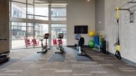 Located at 1201 121st Ave NE in Bellevue, WA, this fitness center is a thoughtfully designed facility that caters to a wide range of workout needs. The space is defined by its dramatic soaring ceilings, which are complemented by large floor-to-ceiling windows along the exterior walls, flooding the interior with an abundance of natural light and offering open views of the surrounding neighborhood. Textured, tile-patterned carpeting covers the floors throughout, providing a durable and functional surface across all workout zones.

The cardio area is generously equipped with treadmills, elliptical machines, stationary bikes, and a rowing machine, all positioned to take advantage of the expansive window views. A dedicated strength training zone features a multi-station cable machine and additional resistance equipment, alongside free weights including kettlebells and medicine balls. TRX suspension trainers are anchored to the walls, offering versatile functional training options.

A large mirrored wall expands the sense of space in the open exercise area, which also features a mounted flat-screen TV. Overhead ceiling fans and linear pendant lighting provide comfort and ambiance. Exposed concrete accent walls and visible ductwork add an industrial character, balancing the modern, clean aesthetic of the overall space.