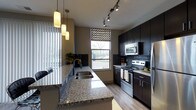 Located at 720 Michigan Ave in Columbus, Ohio, this single-floor residence offers a thoughtfully designed interior with modern finishes throughout. The kitchen is a standout feature, showcasing dark espresso cabinetry that extends to the ceiling, stainless steel appliances including a range and built-in microwave, and a generous granite-topped island with an undermount sink. Pendant lighting and track lighting illuminate the space, while a large window allows natural light to pour in, complementing the wood-look flooring that runs throughout. The kitchen's tile backsplash and contemporary hardware add refined detail to the overall aesthetic. The living area is equally well-appointed, featuring neutral gray walls that serve as a versatile backdrop for the space. Large windows dressed with patterned drapes invite natural light and provide views of the surroundings. The room comfortably accommodates a full seating arrangement, with wood-look flooring continuing the cohesive design language established in the kitchen. The space strikes a balance between modern sophistication and functional livability, with clean lines and a neutral palette that lend themselves to a variety of interior styles. Overall, this residence presents a polished and contemporary living environment in the heart of Columbus.