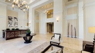 This grand lobby makes an immediate impression with its soaring double-height ceilings, intricate coffered detailing, and expansive polished marble tile flooring that reflects light throughout the space. Large floor-to-ceiling windows flood the entry with natural light, framing views of lush palm trees and the surrounding streetscape, reinforcing a bright and welcoming atmosphere.

At the heart of the lobby, a stately reception desk commands attention, crafted from rich dark wood with ornate leaf-motif metalwork panels and a sleek stone countertop — a striking centerpiece that blends natural and contemporary design. Botanical-inspired artwork adorns the walls above, echoing the organic motifs found throughout the space.

Elegant chandeliers with warm candle-style lighting hang from the lofty ceiling, complemented by understated wall sconces that provide a soft, layered glow. A graceful round entry table anchored by a sculptural dark-wood base and adorned with fresh orchids adds a refined focal point near the entrance.

The lounge seating area features cream upholstered sofas and dark-framed armchairs arranged around a glass-topped coffee table, set upon a classic area rug. Tall decorative sculptures and lush indoor plants complete the composed, resort-like aesthetic of this distinguished lobby.