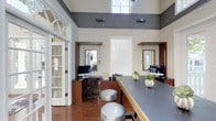 This single-story space showcases a well-appointed business center or leasing office designed with both functionality and aesthetic appeal in mind. The room immediately draws attention to its soaring double-height ceilings, finished in a bold charcoal gray that contrasts sharply with the warm cream-toned walls below. Natural light floods the interior through clerestory windows positioned near the ceiling line, as well as a central white-shuttered window, creating a bright and welcoming atmosphere throughout the day.

The space features rich wood-toned built-in workstations along the perimeter walls, each equipped with desktop computers and framed by recessed mirror panels that add depth and visual interest. A long central communal table with a sleek gray surface runs the length of the room, accompanied by stylish metallic drum stools that provide casual seating. The hardwood flooring in a warm medium tone anchors the space and complements the wood cabinetry throughout.

An elegant French door with arched transom detailing on the left side adds a classical architectural element, balancing the modern furnishings. Subtle pendant lighting and wall-mounted fixtures complete the polished, professional ambiance of this thoughtfully designed workspace.