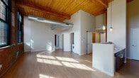 Located at 275 Winchester Ave in New Haven, CT, this single-floor loft-style unit masterfully combines industrial heritage with modern design. The expansive open-plan living area immediately impresses with soaring ceilings clad in warm wood planking supported by heavy timber beams, exposed ductwork, and light-wash hardwood flooring that reflects abundant natural light streaming in throughout the space.

The kitchen is a standout feature, outfitted with sleek, dark wood cabinetry, light stone countertops, stainless steel appliances including a refrigerator and wall oven, and a central island with pendant lighting overhead. Track lighting along the wood-beam ceiling adds both function and industrial flair.

The bedroom area retains the loft's signature character with a full exposed brick wall and large black-framed windows overlooking the surrounding neighborhood. Soft carpeting provides comfort underfoot, and the room connects directly to a generously sized walk-in closet that also features original brick detailing.

The bathroom is thoughtfully designed with tile flooring, accessibility grab bars, and a striking original wooden structural post that echoes the building's historic roots. In-unit laundry is also conveniently included. Throughout the space, the interplay of reclaimed materials and contemporary finishes creates a distinctive, urban ambiance.