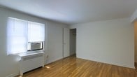 This single-floor space presents a clean and minimalist interior defined by warm hardwood flooring that runs throughout, lending a classic and timeless character to the room. The walls are finished in a neutral white, creating a bright and airy atmosphere that maximizes the sense of openness within the space.

Natural light enters through a window equipped with horizontal blinds, beneath which sits a window-mounted air conditioning unit, providing climate control during warmer months. A cast-iron radiator beneath the window serves as the primary heat source, a practical feature well-suited for cooler seasons.

An open closet with a visible hanging rod offers convenient built-in storage, adding functional value to the room without requiring additional furniture. The door to the closet is a standard white panel design, consistent with the overall clean aesthetic of the space.

The room is straightforward in layout, with a simple, unadorned ceiling and minimal architectural detailing. The overall ambiance is functional and understated, presenting a blank canvas well-suited for personalization. The combination of hardwood flooring, neutral tones, and practical built-in features gives the space a no-frills, utilitarian appeal.