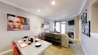 This single-floor space offers an open-concept layout that seamlessly integrates the dining and living areas into one cohesive environment. The dining area is anchored by a modern concrete-style table surrounded by vibrant orange accent chairs, set with place settings that add a pop of color against the neutral backdrop. A sleek black console table serves as a stylish divider between the two zones, topped with decorative accessories.

The living area features a plush sofa accented with colorful throw pillows, a patterned area rug, and a wall-mounted flat-screen television positioned above a classic white fireplace with a traditional mantle — a standout focal point in the room. Natural light filters in through a frosted glass door, brightening the space alongside recessed ceiling lighting.

The walls are painted in a soft gray tone and adorned with an eclectic gallery wall, complemented by a large, bold pop-art canvas that adds personality and visual interest. Crown molding along the ceiling adds a refined architectural touch. Overall, the space balances contemporary design with vibrant, artistic elements.