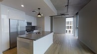 This contemporary single-floor space showcases an open-concept layout that seamlessly connects the kitchen and living areas, creating a bright and functional environment. The kitchen is a standout feature, equipped with high-gloss white cabinetry, a dark quartz waterfall island with an integrated sink, and a full suite of stainless steel appliances including a refrigerator, microwave, and range. An industrial-style pendant light hangs above the island, adding a warm focal point to the otherwise clean, minimalist aesthetic. The exposed ceiling with visible ductwork and structural elements lends an urban, loft-inspired character to the space. Light-toned hardwood flooring runs throughout, contributing to a sense of warmth and continuity. A floor-to-ceiling window wall on one end of the living area allows abundant natural light to pour in, framing views of the surrounding urban environment. A barn-style sliding door track is visible, suggesting a flexible room division option. The overall design balances modern finishes with industrial touches, making for a stylish and versatile space well-suited to contemporary living.