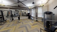 Located at 430 Allied Pl in Gaithersburg, Maryland, this well-appointed fitness center offers a comprehensive workout environment across a single, open floor. The space is designed with both function and aesthetics in mind, featuring a distinctive herringbone-patterned carpet in warm gold and grey tones that lends a polished, contemporary feel underfoot.

The facility is outfitted with a wide array of equipment to support diverse training needs. A cable crossover machine and smith/squat rack serve as centerpieces for strength training, while a full dumbbell rack stocked with a range of weights, kettlebells, and weight plates accommodate free weight workouts. Multiple selectorized resistance machines line the walls for targeted muscle training, and a flat utility bench supports a variety of exercises.

Large wall-mounted mirrors span one side of the room, visually expanding the space and allowing users to monitor their form during workouts. Elegant pendant-style drum light fixtures provide warm, even illumination throughout. A wall-mounted flat-screen television adds an entertainment element to the space.

Natural light filters in through multiple windows dressed with horizontal blinds, offering views of the surrounding greenery. Exposed ductwork along the ceiling adds an industrial accent that complements the neutral grey walls. The overall ambiance is clean, functional, and inviting for a focused fitness experience.