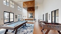 Located at 1181 Manhattan Blvd in Dayton, Kentucky, this single-floor community lounge and game room is a standout amenity space designed for both recreation and socializing. The room immediately impresses with its dramatically soaring ceilings clad in warm wood paneling, which contrast beautifully against the crisp white walls and create a striking architectural focal point. A geometric linear pendant light fixture anchors the central game area, where a full-size pool table takes center stage atop a bold blue abstract area rug. Alongside it, a solid wood shuffleboard table and a foosball table provide additional entertainment options. Toward one end of the room, a sleek white bar counter lined with cognac-colored leather barstools sits beneath a wood-paneled accent wall, complete with a mounted TV, pendant lighting, and a curated display of barware. A casual dining table with modern chairs offers additional seating nearby. On the opposite end, a lounge area with comfortable seating and a mounted flat-screen TV provides a relaxing retreat. Warm hardwood flooring runs throughout, while steel-framed multi-pane windows flood the space with natural light and offer views to the surrounding community. The overall ambiance is modern, warm, and inviting.