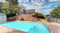 This inviting outdoor amenity space centers around a generously sized swimming pool with crystal-clear blue water, surrounded by a broad tiled sun deck designed for both relaxation and socializing. The deck is well-furnished with lounge chairs, tables, and shade umbrellas, providing ample options for sunbathing or casual outdoor dining. A small, dedicated pool house structure sits poolside, likely housing equipment and safety information for guests.

Framing the pool area is a sturdy black wrought iron fence that encloses the space while maintaining open sightlines to the surrounding tree-lined grounds. The mature trees provide natural shade and contribute to a serene, park-like atmosphere throughout the area.

Set prominently above the pool deck is a single-story clubhouse building featuring a pitched shingle roof, light gray siding, and a warm wood-toned elevated deck with decorative railings and a staircase connecting it directly to the pool level. The building's elevated position offers an excellent vantage point over the pool and surrounding landscape, adding a distinctive architectural focal point to the overall setting. The combination of natural stone retaining walls, rich wood deck accents, and lush greenery creates a cohesive, resort-style ambiance that is both functional and aesthetically appealing.