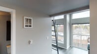 This two-floor unit offers a thoughtfully organized layout spread across two distinct levels. The main level features hard surface flooring throughout and is anchored by a compact kitchen equipped with white cabinetry, light countertops, a stainless steel sink, and an over-the-range microwave — providing efficient functionality in a clean, minimal design. A full bathroom is conveniently located on the main floor, featuring a vanity with dark cabinetry, a wall-mounted mirror with integrated lighting, and a toilet, all finished in a neutral palette. Wall-mounted heating units are present throughout, ensuring climate control across the space. Large floor-to-ceiling windows on the main level flood the interior with natural light and offer views of the surrounding urban environment. Access to the upper loft level is provided via a ladder-style staircase, adding a distinctive architectural element. The upper level is carpeted and provides an open, flexible area well-suited for use as a sleeping or lounge space. A portable air conditioning unit is also present on this level. A separate carpeted room with a wall-mounted shelf offers additional storage or flexible use. The overall aesthetic is clean and modern, characterized by neutral tones, simple finishes, and efficient use of space throughout both floors.