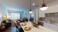 This single-floor residence offers a thoughtfully designed open-concept layout that seamlessly connects the kitchen, dining, and living areas. The kitchen is a standout feature, outfitted with crisp white shaker cabinetry, a gray subway tile backsplash, stainless steel appliances including a refrigerator, range, and built-in microwave, and a generous island topped with quartz countertops. Pendant lighting above the island adds a modern touch, while dark hardwood-style flooring anchors the space with warmth and continuity.

The living area is bright and inviting, benefiting from large windows dressed with blinds and curtains that allow natural light to fill the room. Two well-proportioned bedrooms provide comfortable retreats, each featuring plush carpeting, ample natural light, and thoughtful design details. One bedroom includes a walk-in closet equipped with multi-tiered wire shelving, offering generous storage capacity.

The bathroom is finished in a clean, contemporary style with white cabinetry, a wide vanity mirror with strip lighting, dark hardwood-style flooring, and a glass-enclosed shower. Throughout the residence, a neutral color palette of soft whites and grays creates a cohesive and calm ambiance, enhanced by consistent trim and door detailing that lend a polished, refined character to the overall space.
