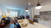 This single-floor residence offers a thoughtfully designed open-concept layout that seamlessly connects the kitchen, dining, and living areas. The kitchen is a standout feature, outfitted with crisp white shaker cabinetry, a gray subway tile backsplash, stainless steel appliances including a refrigerator, range, and built-in microwave, and a generous island topped with quartz countertops. Pendant lighting above the island adds a modern touch, while dark hardwood-style flooring anchors the space with warmth and continuity.

The living area is bright and inviting, benefiting from large windows dressed with blinds and curtains that allow natural light to fill the room. Two well-proportioned bedrooms provide comfortable retreats, each featuring plush carpeting, ample natural light, and thoughtful design details. One bedroom includes a walk-in closet equipped with multi-tiered wire shelving, offering generous storage capacity.

The bathroom is finished in a clean, contemporary style with white cabinetry, a wide vanity mirror with strip lighting, dark hardwood-style flooring, and a glass-enclosed shower. Throughout the residence, a neutral color palette of soft whites and grays creates a cohesive and calm ambiance, enhanced by consistent trim and door detailing that lend a polished, refined character to the overall space.