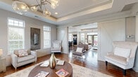 Located at 4170 Green Clover Drive in Hilliard, Ohio, this thoughtfully designed single-floor space offers a seamless blend of sophistication and comfort. The entrance welcomes visitors with rich hardwood flooring and a clean, open aesthetic that flows naturally into the main living areas.

The living room is a standout feature, distinguished by tray ceilings that add architectural depth and dimension. A striking modern chandelier with globe accents serves as a focal point, casting warm light across the space. Multiple large windows fitted with plantation shutters line the walls, inviting generous natural light while maintaining a sense of privacy. The warm hardwood floors extend throughout, grounding the space with timeless character.

A secondary lounge area is defined by decorative white columns framing a bold, patterned accent wall, creating an intimate and visually dramatic nook. This area transitions smoothly into an additional seating zone equipped with a large wall-mounted television, sleek bar-height seating, and a marble-top coffee table — ideal for both relaxation and entertainment.

The entrance area opens directly to the exterior via French doors with transom windows above, reinforcing the bright, airy atmosphere. Tray ceiling detailing continues here as well, lending a cohesive architectural language throughout. The overall ambiance is polished and inviting, with a neutral palette, upscale finishes, and thoughtful design details at every turn.