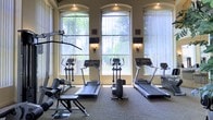 This fitness center offers a thoughtfully designed workout space that balances functionality with an inviting aesthetic. The room is anchored by a row of large, arched windows fitted with vertical blinds, allowing generous natural light to fill the space while providing views of lush outdoor greenery. The warm, neutral-toned walls are complemented by decorative wall sconces that add an elegant touch to the environment.

The cardio section is well-stocked, featuring multiple treadmills and elliptical trainers positioned to take advantage of the natural light and garden views — ideal for an energizing workout experience. A multi-function strength training cable machine and a dedicated weight bench provide options for resistance and strength training, catering to a variety of fitness routines.

Recessed ceiling lighting supplements the natural light, ensuring the space remains well-lit at all hours. The carpeted flooring adds a degree of comfort underfoot while contributing to the room's polished appearance. Wall-mounted televisions are positioned throughout the room, allowing users to stay entertained during their workouts.

Overall, this fitness center combines practical equipment with refined architectural details, creating a comfortable and motivating environment suited for a broad range of fitness activities.