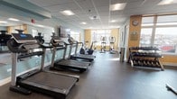This fitness center is a thoughtfully designed, single-level space that accommodates a comprehensive range of workout activities. The layout is organized into distinct functional zones, allowing users to move efficiently between cardio, strength, and free-weight training areas.

The cardio section lines one side of the room and features multiple treadmills, upright and recumbent stationary bikes, and elliptical machines. Wall-mounted flat-screen televisions are positioned throughout for entertainment during workouts. A large wall mirror expands the visual depth of the cardio area, while a striking full-wall photographic mural of a bridge perspective serves as a bold design accent.

The strength and free-weight zone includes a multi-function cable machine, a flat utility bench, a tiered dumbbell rack with a full set of weights, and additional resistance equipment. Stability balls add versatility to the training options available.

The flooring throughout is durable rubber gym matting, appropriate for high-traffic fitness use. The drop ceiling features recessed panel lighting supplemented by flush-mounted fixtures, providing consistent, even illumination. Warm amber and soft teal wall colors create an energizing yet approachable atmosphere. Windows along one wall admit natural light and offer exterior views. Motivational artwork and a wall clock complete the interior details of this well-appointed fitness facility.