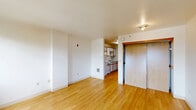 Located at 1212 SW Clay St in Portland, Oregon, this efficiently designed single-floor unit makes smart use of its compact footprint. The main living area is bright and open, featuring warm hardwood flooring that extends throughout the space, crisp white walls, and clean lines that give the unit an airy, unfussy feel. A pair of windows along one wall frames views of the surrounding Portland skyline and mature trees, allowing natural light to fill the room. A sliding wood-panel closet door adds a touch of warmth and character to the living space, complemented by a second closet providing additional storage. The galley-style kitchen is neatly tucked adjacent to the main living area and is equipped with white cabinetry featuring wood trim, an electric range, refrigerator, and a ventilation hood. A pass-through view from the kitchen back into the living area maintains an open, connected feel. The bathroom includes a large vanity with ample counter space, a wide mirror, and white cabinetry consistent with the kitchen's design language. Hardwood flooring continues seamlessly throughout, tying the entire unit together in a cohesive, practical aesthetic well-suited to urban living in Portland.