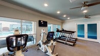 This fitness center is a thoughtfully designed, single-floor exercise facility offering a comprehensive range of workout equipment and functional spaces. The room is finished with crown molding along the teal-painted ceiling, recessed lighting, and dual ceiling fans that maintain air circulation throughout the space. Neutral-toned carpet covers the floor, providing a comfortable surface underfoot.

The cardio area features multiple Spirit-brand treadmills arranged in a row, positioned along large sliding glass windows that overlook an outdoor pool area, allowing natural light to fill the space and offering pleasant views during workouts. A wall-mounted flat-screen television is positioned nearby for entertainment. Stationary bikes and rowing equipment are also available in this section.

A separate zone accommodates strength and flexibility training, housing a resistance band station, a weight bench, a dumbbell rack, and additional cable equipment. This area is partially enclosed by a floor-to-ceiling mirrored glass partition wall, creating a distinct training space while maintaining visual openness.

Access to adjoining areas is provided through both frosted-glass French doors and a single frosted-glass interior door, contributing to the overall clean, polished aesthetic of the facility.