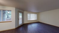 Located at 1616 NE 16th Way in Gresham, Oregon, this single-floor residence offers a well-organized layout with a blend of functional design and comfortable living spaces. The main living area is defined by rich, dark hardwood flooring that extends throughout the common areas, complemented by neutral cream-toned walls and wood-trimmed doorways and baseboards. A large front-facing window fitted with vertical blinds and a door with window panel allow natural light to fill the living room, creating a bright and inviting atmosphere. A ceiling fan adds practical comfort to the space. The kitchen features an open-concept design anchored by a peninsula counter that separates it from the living area. White cabinetry with wood-accented trim lines the walls, offering ample storage, while appliances include a full-size refrigerator, electric range, microwave, and dishwasher. The adjacent bathroom is well-appointed with a wood-framed vanity, wide mirror, soaking bathtub with shower, and dark hardwood flooring. The bedrooms are carpeted in neutral tones and offer generous proportions, with at least one bedroom benefiting from a window with outdoor views and access to a walk-in closet. The textured ceilings throughout contribute to the home's classic residential character.