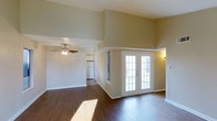 Located in Arlington, this single-floor space impresses with its dramatic vaulted ceilings that create an expansive, airy atmosphere throughout the main living area. Rich, dark hardwood-style flooring runs seamlessly across the space, adding warmth and a polished aesthetic to the interior. A standout feature is the set of classic French doors, framed in crisp white, which invite abundant natural light and offer a refined connection to the outdoors. The open layout of the main living area is enhanced by a ceiling fan, providing both comfort and a functional design touch. The space transitions naturally into a kitchen area, visible in the background, which features white cabinetry and appliances, maintaining a clean and cohesive color palette throughout. The warm, neutral tones of the walls complement the flooring and trim, creating a cohesive and inviting ambiance. The combination of high ceilings, natural light, and open-concept flow makes this Arlington space well-suited for both everyday living and entertaining.