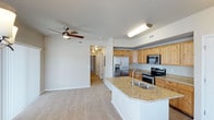 This single-floor residence showcases an open-concept layout that seamlessly connects the kitchen and living area, creating a spacious and functional environment. The kitchen is a standout feature, equipped with stainless steel appliances including a French door refrigerator with an in-door ice and water dispenser, an electric range, and a built-in microwave. Natural-toned wood cabinetry provides ample storage, complemented by granite countertops that extend to a peninsula island complete with a double-basin sink — ideal for meal prep and casual dining. The living area flows naturally from the kitchen, featuring neutral carpeting and a ceiling fan that adds both comfort and airflow. A hallway visible from the main living space leads to additional rooms, suggesting a well-organized floor plan with private quarters separated from the common areas. Throughout the residence, white walls and neutral tones create a clean, cohesive aesthetic, while consistent ceiling fixtures provide practical lighting. The overall ambiance is modern yet inviting, with quality finishes that balance style and everyday functionality.