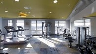 Located at 3001 Village Market Pl in Morrisville, NC, this fitness center offers a well-appointed, single-floor space designed to support a comprehensive workout experience. The room features a bold yellow ceiling accented by recessed lighting, creating a bright and energizing atmosphere that complements the abundant natural light streaming in through large floor-to-ceiling windows along the perimeter walls. These expansive windows provide open views of the surrounding area, contributing to an airy, motivating environment. The space is equipped with a thoughtfully curated selection of exercise machines, including a cable crossover station, strength training equipment, and cardio machines such as a treadmill. The flooring consists of a patterned, multi-toned carpet that adds visual interest while providing comfort underfoot. Mirrored walls further enhance the sense of openness and allow users to monitor their form during workouts. The neutral gray walls, combined with the vivid ceiling and natural light, strike a balance between functional design and modern aesthetics, making this fitness center a well-rounded facility for residents and visitors in the Village Market Place community.